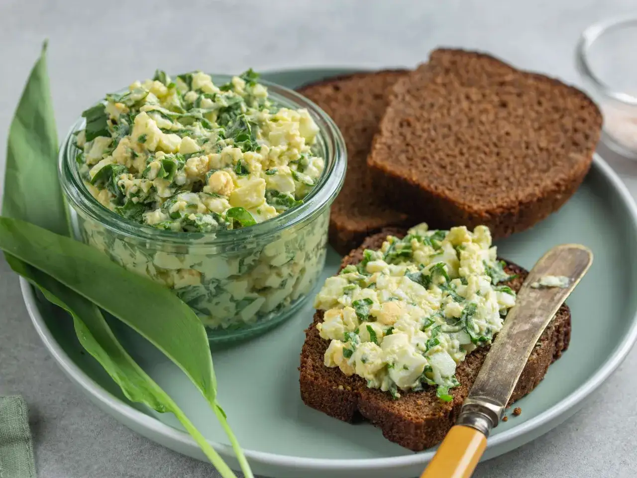 Ein Glas mit cremigem Eiersalat, verfeinert mit frischen Kr&auml;utern, daneben eine Scheibe dunkles Brot mit dem Eiersalat. Ein einfaches eiersalat rezept.