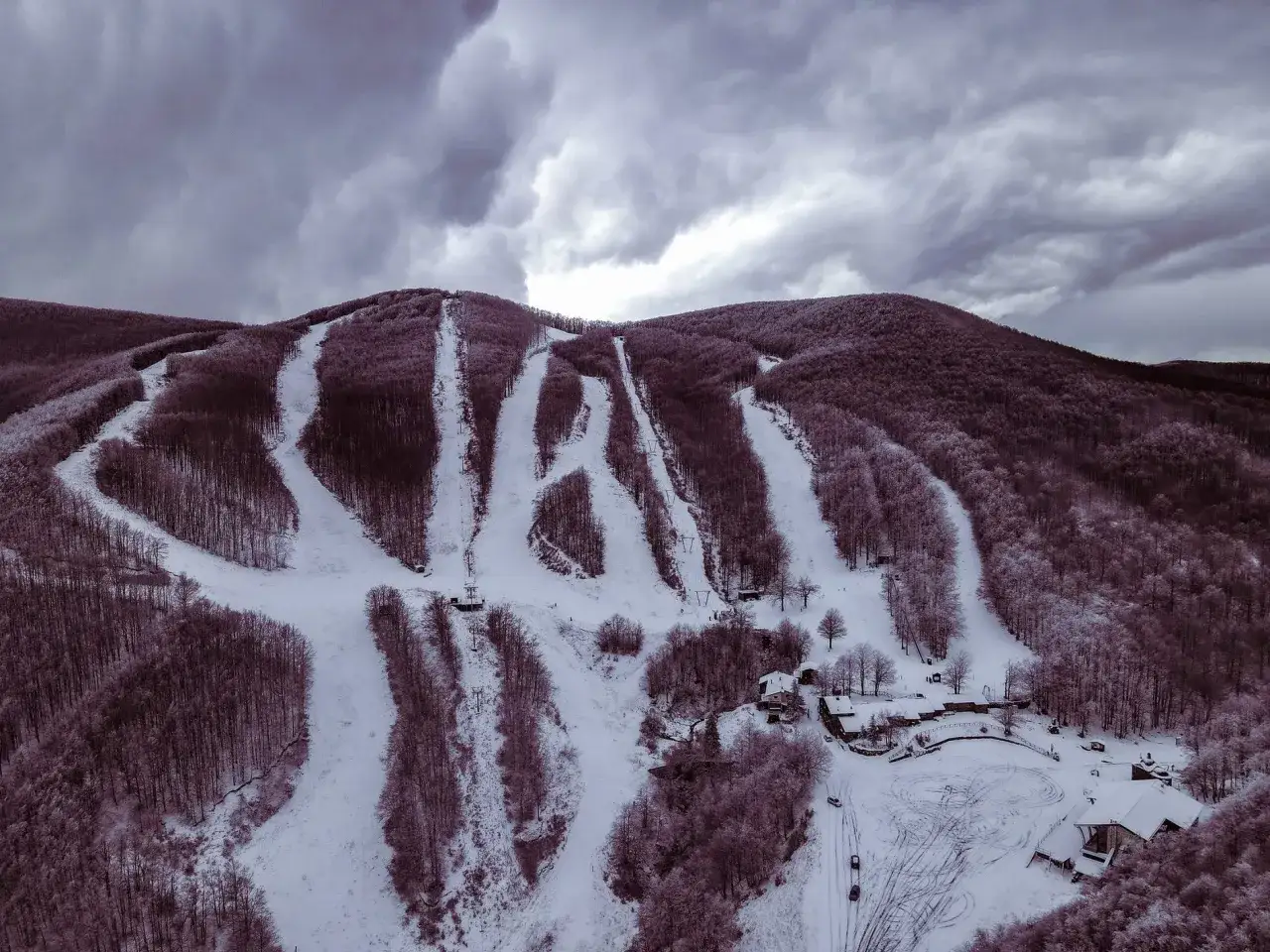 Piste innevate e alberi spogli sul monte zum Zeri, sotto un cielo minaccioso. Un piccolo complesso di edifici si intravede tra gli alberi.