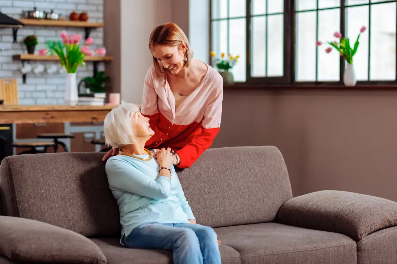 Wsparcie i troska w centrum rehabilitacyjno opiekuńczym dla osób przewlekle chorych somatycznie. Młoda kobieta pochyla się nad starszą panią siedzącą na kanapie.
