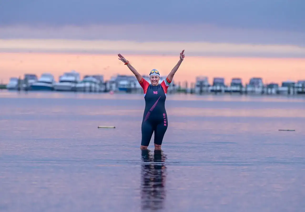 A triumphant swimmer in a wetsuit raises arms in victory, a testament to how to train for an Ironman. Boats line the horizon at dawn.