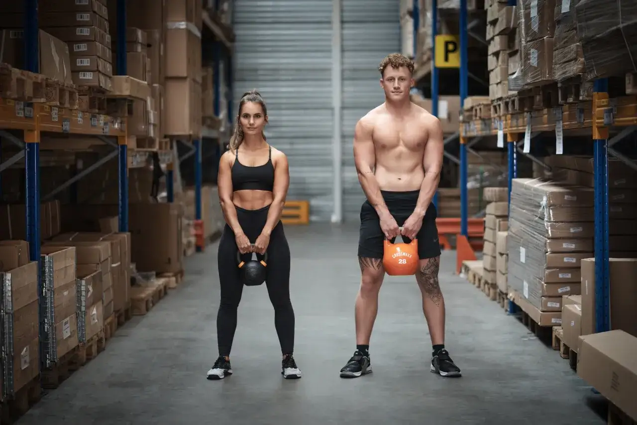 Ein Mann und eine Frau mit Kettlebells in einem Lagerhaus, bereit f&uuml;r ihren HIIT Trainingsplan im Fitnessstudio.
