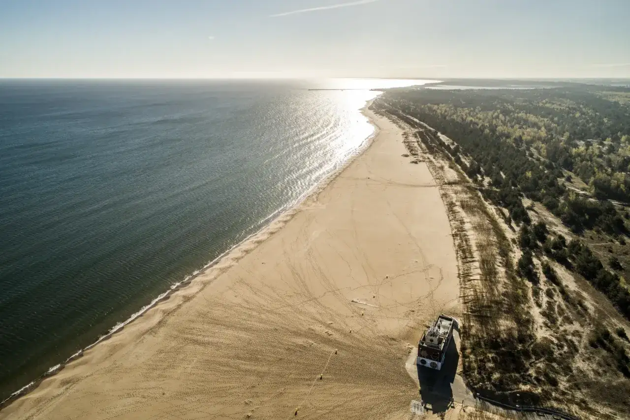 Plaża nudyst&oacute;w stogi. Szeroka, piaszczysta plaża nad Bałtykiem, z lasem sosnowym w tle. Słońce odbija się od wody.