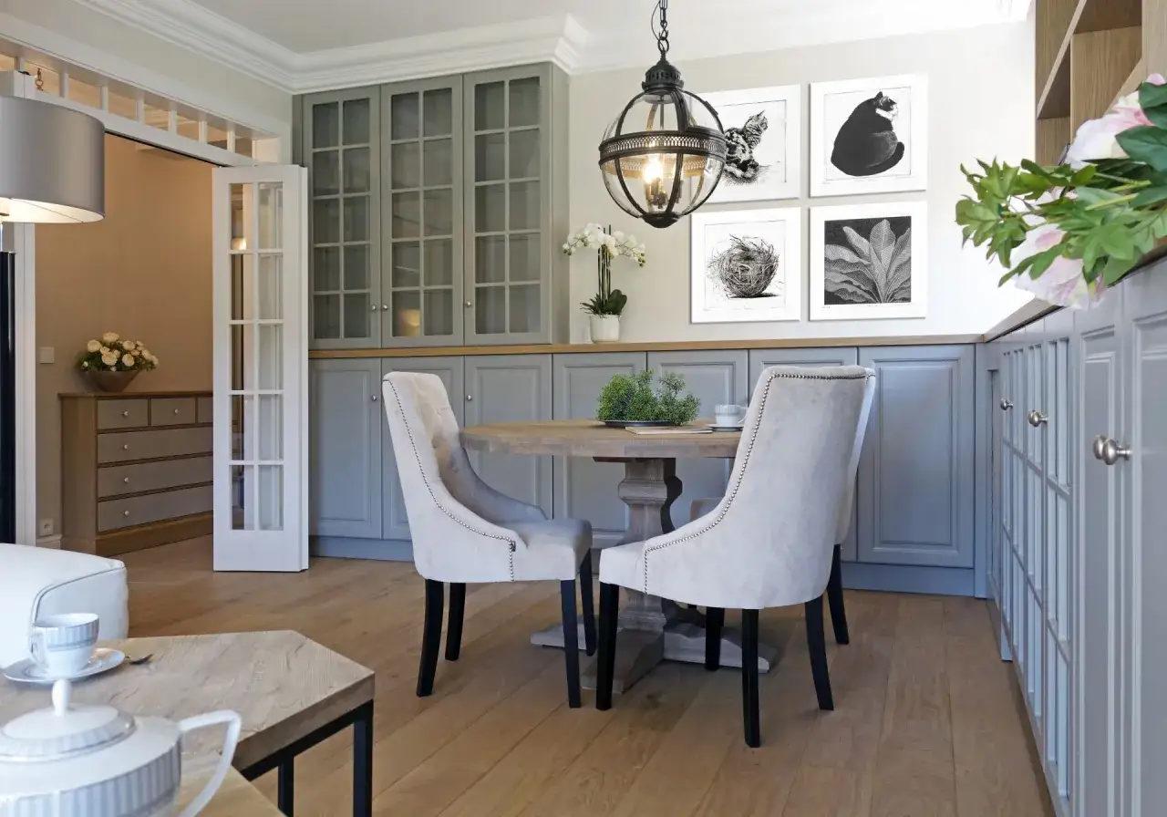 Eleganckie wnętrza w stylu hamptons: okrągły stół, tapicerowane krzesła, szare meble kuchenne i grafiki na ścianie.