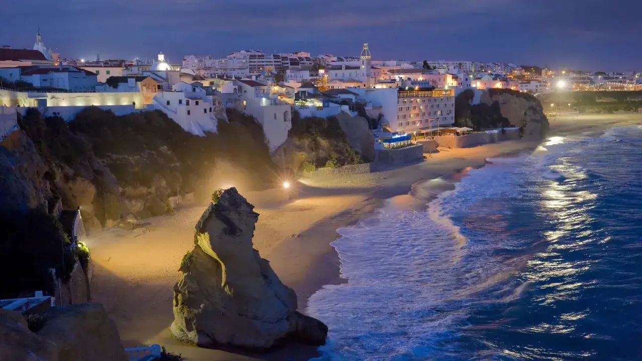 Nachtansicht von Albufeira an der Algarve. Wei&szlig;e H&auml;user auf Klippen, beleuchteter Strand mit Felsen und Wellen.