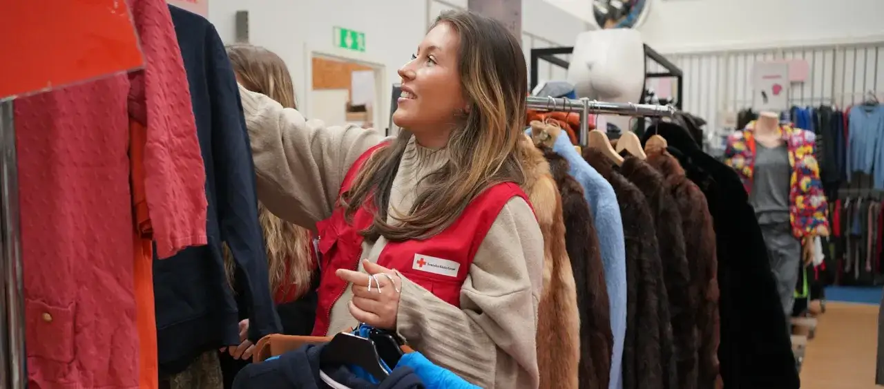 En kvinna i röd väst från Röda Korset sorterar kläder i en butik. Kanske hon letar efter myrorna lediga jobb här?