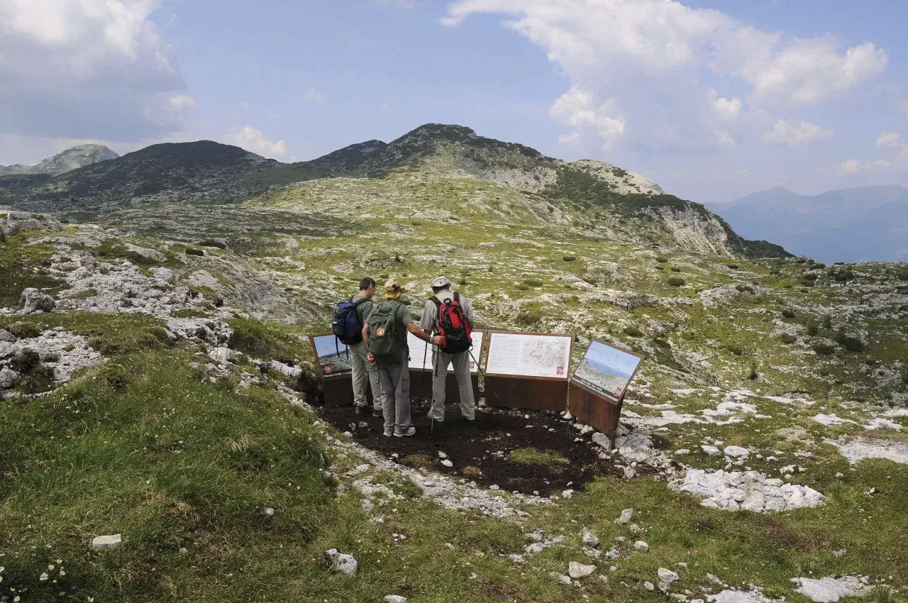 Escursionisti leggono pannelli informativi sul **monte Ortigara come arrivare**. Paesaggio montano roccioso e verdeggiante.