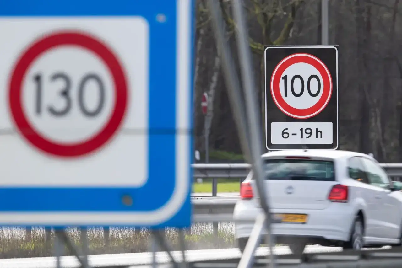 Znaki drogowe na autostradzie w Holandii: ograniczenie do 130 km/h i 100 km/h w godzinach 6-19. Biały samochód jedzie po drodze.