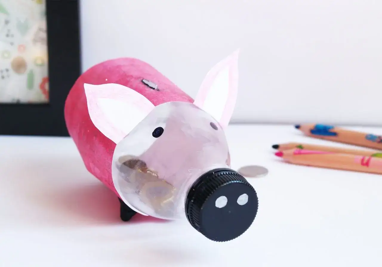 Urocza skarbonka diy z butelki, ozdobiona r&oacute;żowym papierem i uszkami. W jej brzuszku widać monety.
