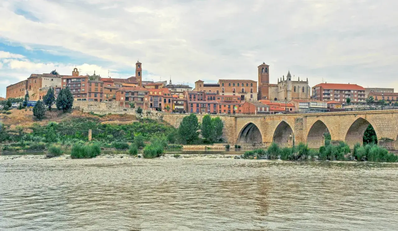 Un puente de piedra cruza un río frente a un pueblo con tejados rojos y torres. ¡Un ejemplo de los pueblos bonitos cerca de Valladolid!