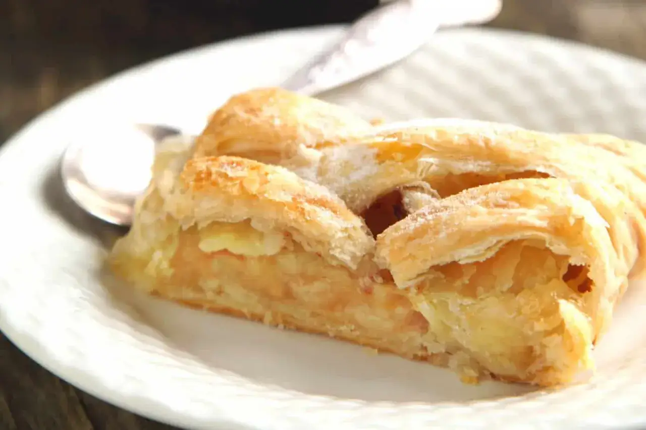 Un trozo de strudel de manzana dorado y hojaldrado, espolvoreado con azúcar glas, servido en un plato blanco con una cuchara.