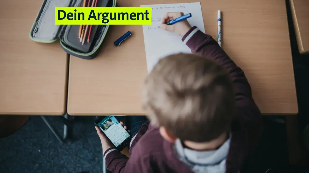 Schüler spickt auf dem Handy, während er im Unterricht sitzt. Stifte und Heft liegen bereit.