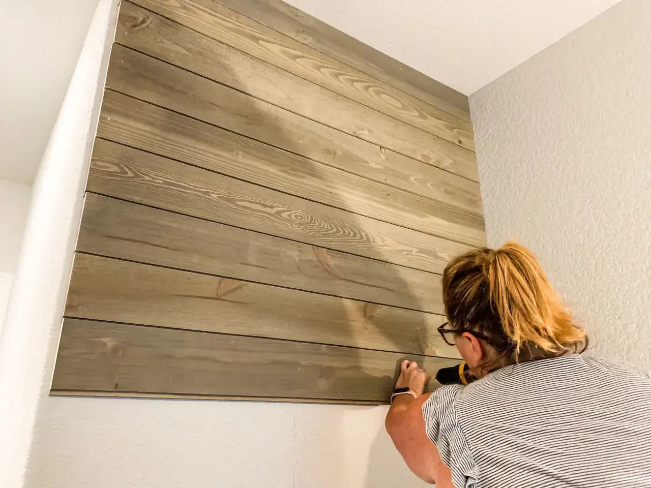 A woman demonstrates how to install shiplap, drilling into the wooden planks on a wall.