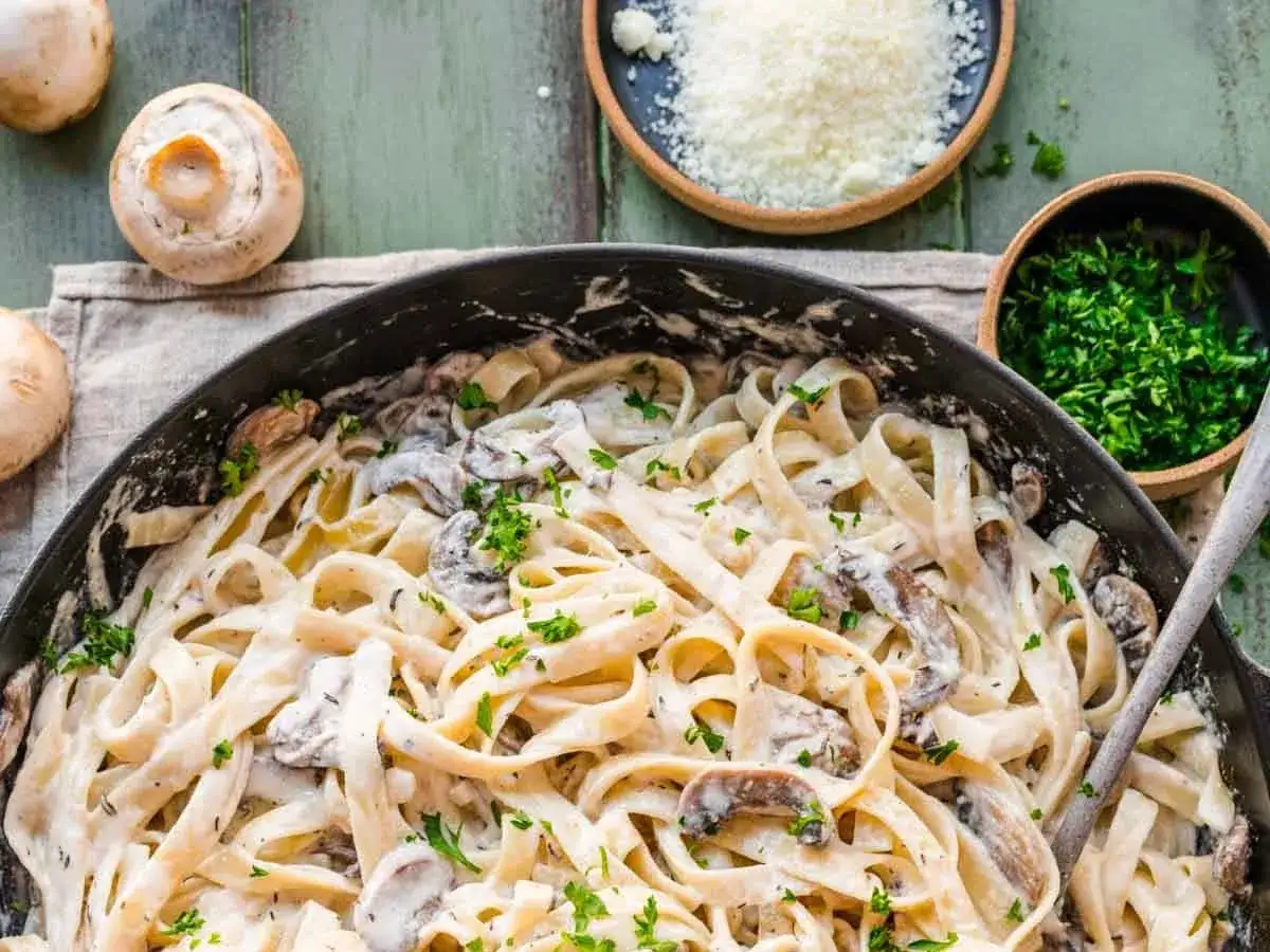 Cremige Pasta mit Pilzen, garniert mit Petersilie. Daneben Schüsselchen mit geriebenem Parmesan und gehackter Petersilie.
