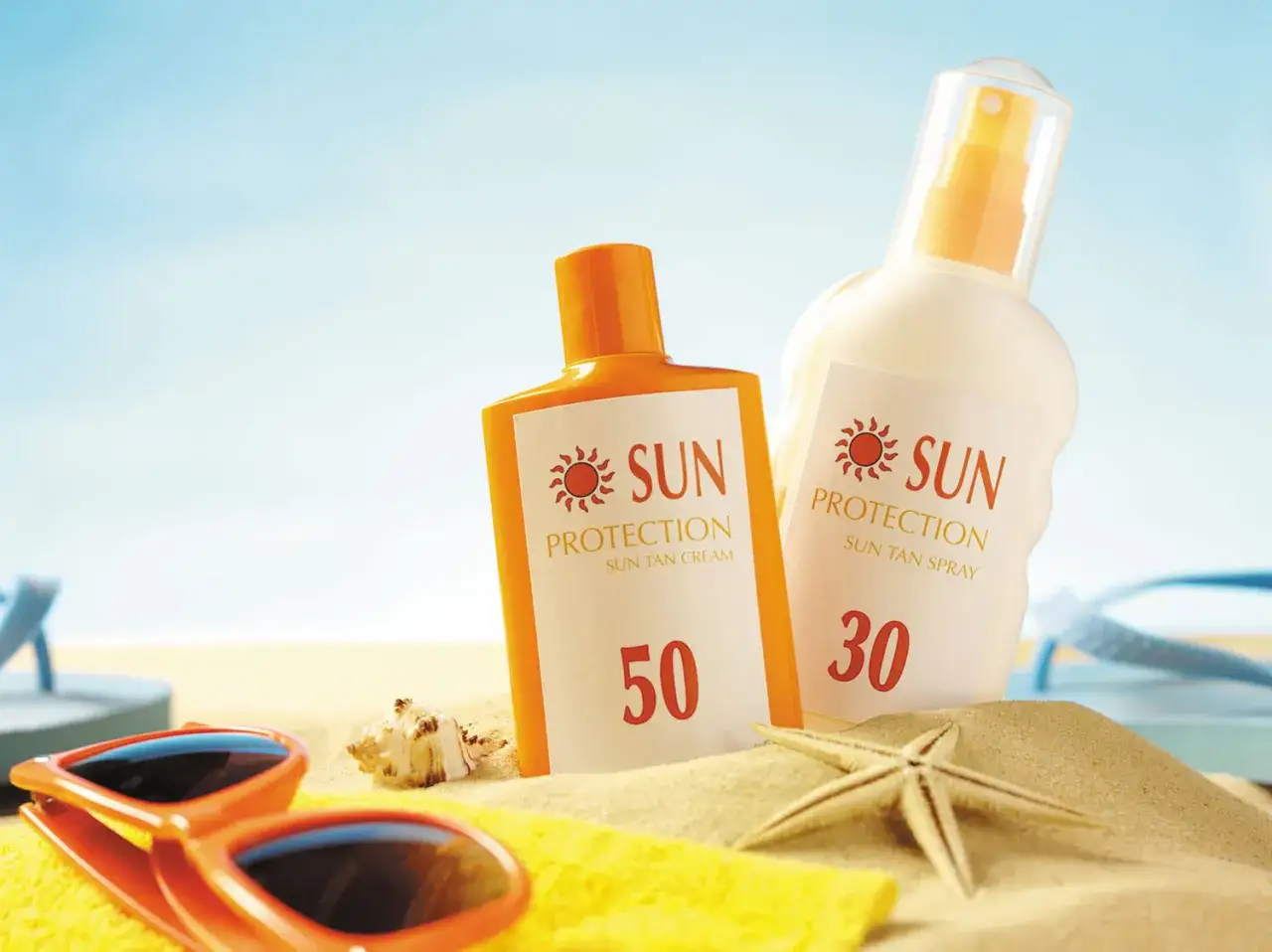 Krem z filtrem SPF 50 chroni sk&oacute;rę przed słońcem. Na plaży leżą okulary, muszelki i dwa kosmetyki do opalania.