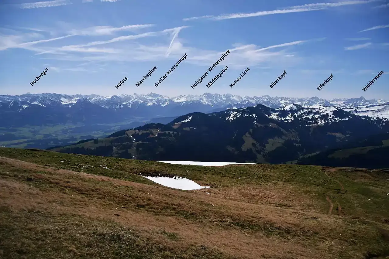Blick &uuml;ber gr&uuml;ne H&uuml;gel und schneebedeckte Gipfel. Ein Pfad schl&auml;ngelt sich den Hang hinauf, wie eine Leiter zum Steineberg.