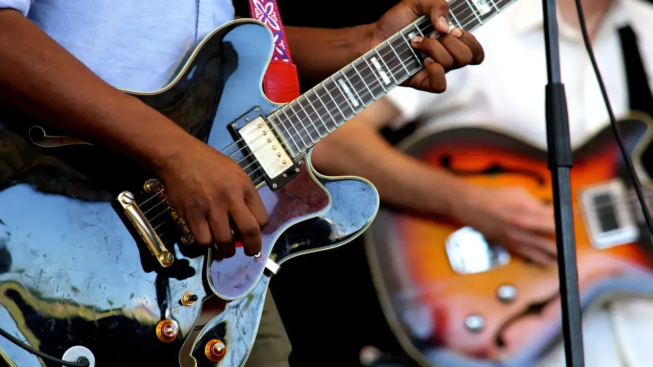 Hände spielen Gitarre, während im Hintergrund eine weitere Gitarre und ein Mikrofonständer zu sehen sind. Die Musik ist so gut, dass man fast die **Band Namen** erraten kann.
