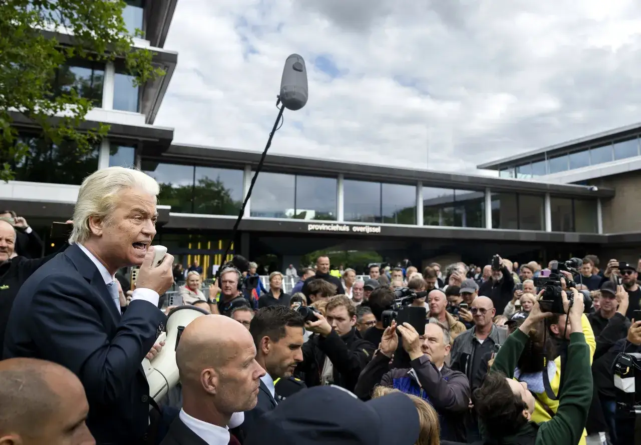 Een politicus spreekt met een megafoon tot een menigte, omringd door journalisten met camera's. Dit toont de kern van wat is populisme: directe communicatie met het volk.