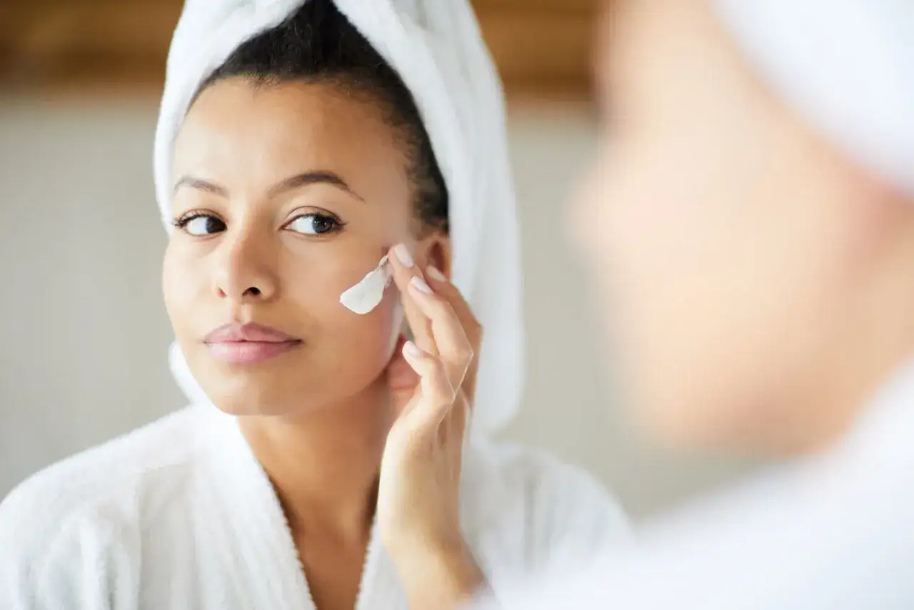 Donna con turbante e accappatoio si applica crema sul viso. Forse mettere la crema tutti i giorni fa male?
