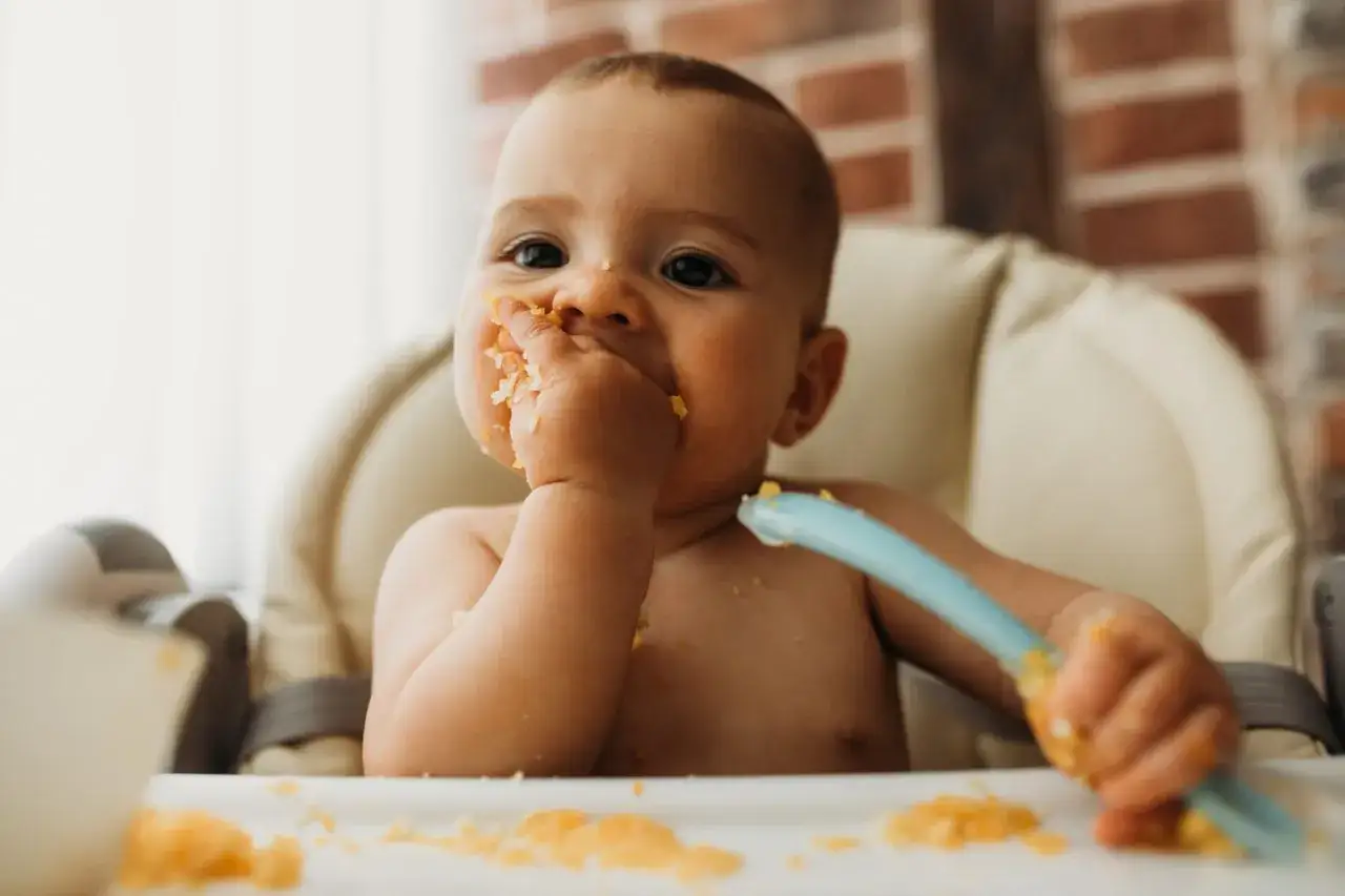 Kleines Baby mit Hummus im Gesicht und auf den Händen, das versucht, sich selbst zu füttern.