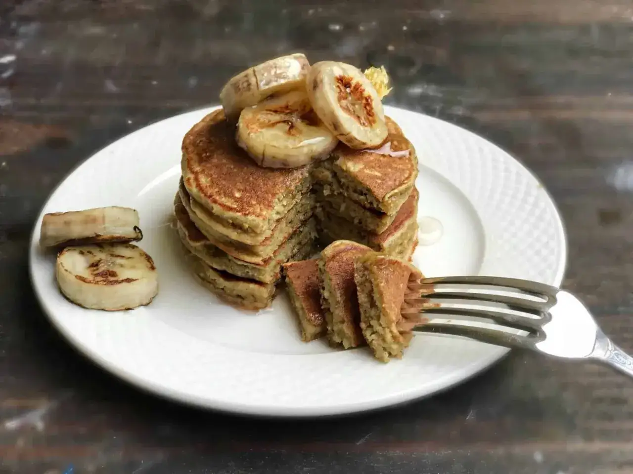 Stos puszystych, trzyskładnikowych placków bananowych, polanych syropem i ozdobionych plasterkami karmelizowanego banana. Widelec nabiera kawałek placka.