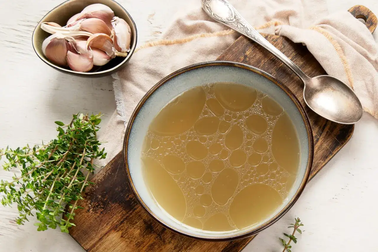 Un bol de bouillon de poulet, des gousses d'ail et du thym frais sur une planche en bois.