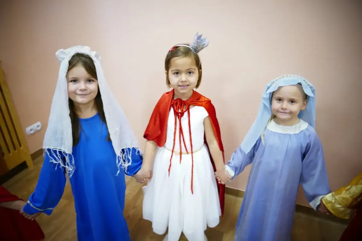Trzy dziewczynki w strojach świętych trzymają się za ręce. Jedna w niebieskiej sukni i białym welonie, druga w białej sukni i czerwonym płaszczyku, trzecia w błękitnej sukni i welonie.