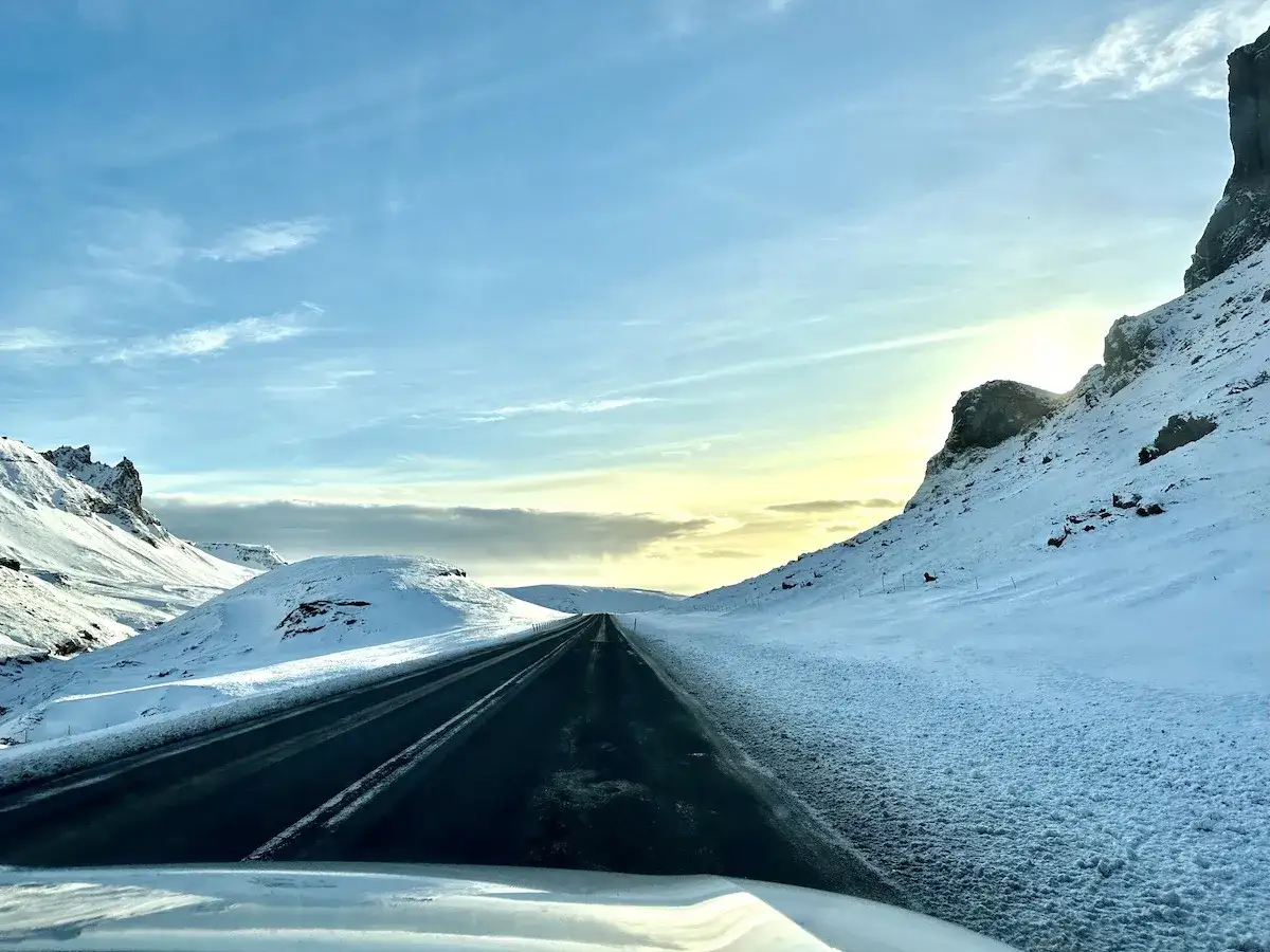 Driving in Iceland in December, a dark asphalt road cuts through a snow-covered valley under a bright, cloudy sky.
