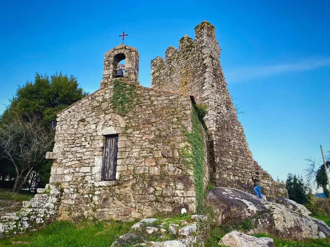 Iglesia de piedra antigua en Catoira, Galicia, con un campanario y una torre defensiva. Un fot&oacute;grafo captura la escena.