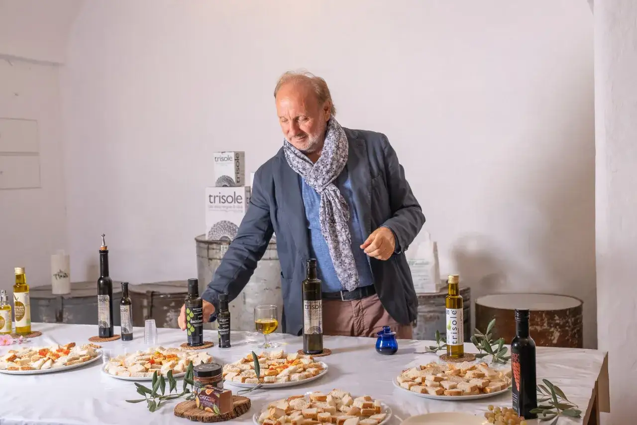 Uomo con sciarpa presenta una degustazione di olio evo, con bottiglie di trisole e crostini su un tavolo.