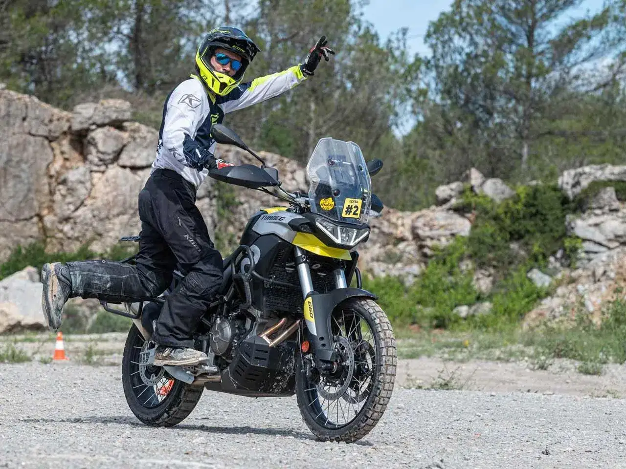 Motociclista con casco amarillo saluda sobre una moto trail, lista para la aventura en terreno rocoso.