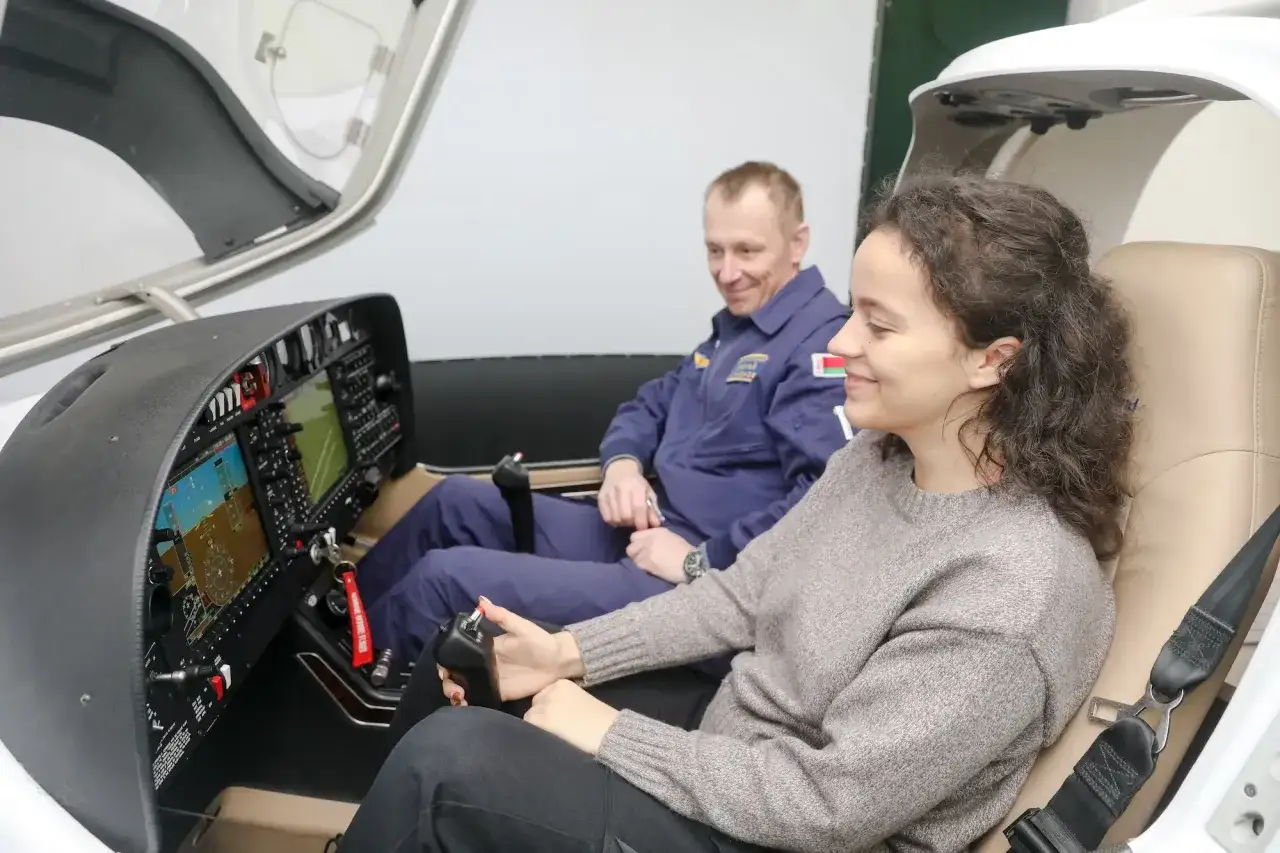 Uśmiechnięta dziewczyna trzyma drążek sterowy, a obok niej siedzi pilot samolotu w niebieskim kombinezonie.