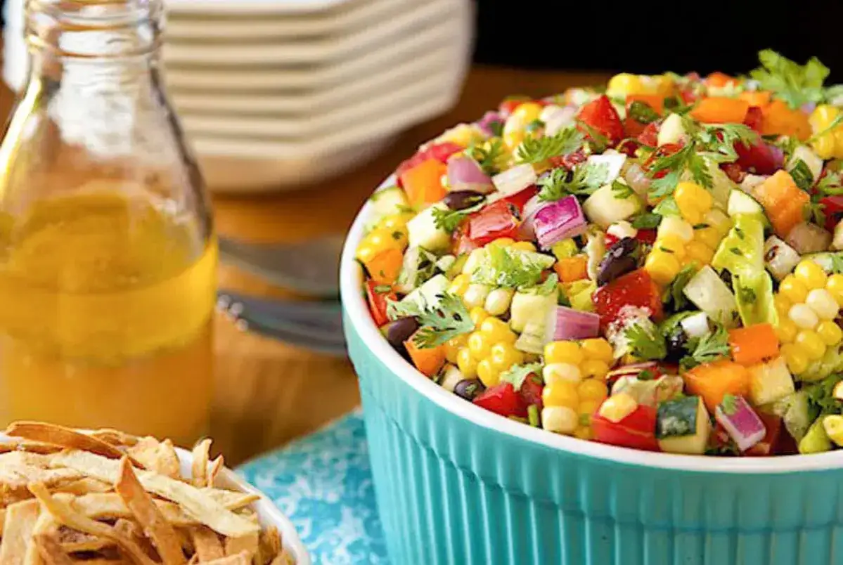 Frischer, bunter mexikanischer Salat mit Mais, Bohnen, Tomaten und Koriander in einer t&uuml;rkisfarbenen Sch&uuml;ssel, daneben eine Flasche Dressing und Tortilla-Streifen.