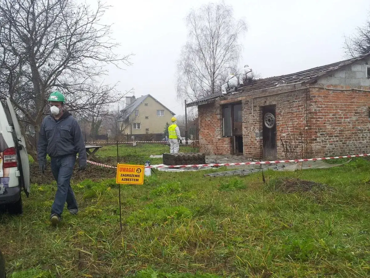 Pracownicy w kombinezonach usuwają eternit z dachu. Tablica ostrzega: 