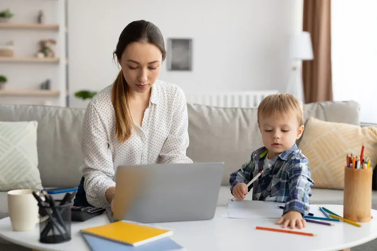 Kobieta pracuje przy laptopie, podczas gdy jej zdrowy syn rysuje obok. Opieka nad dzieckiem i praca w domu.