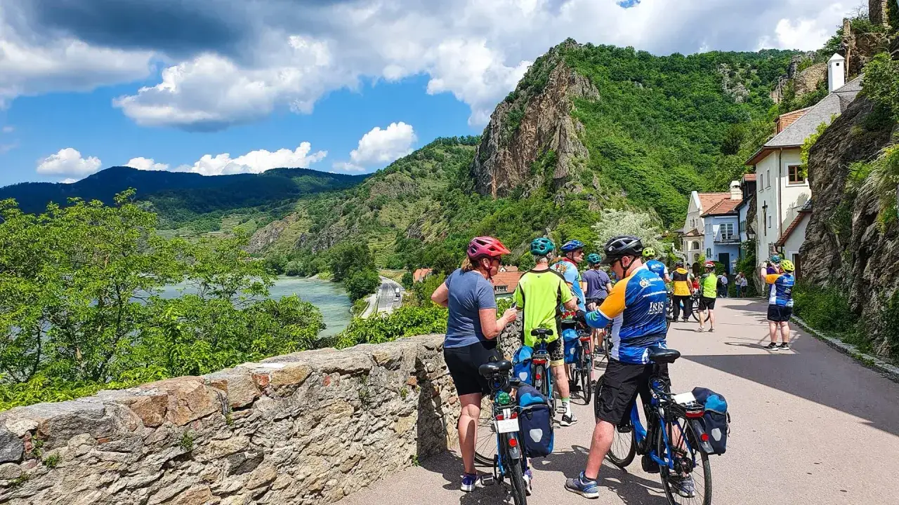 Grupo de ciclistas disfrutando de un paseo por el Danubio en bici, con un paisaje monta&ntilde;oso y casas pintorescas.