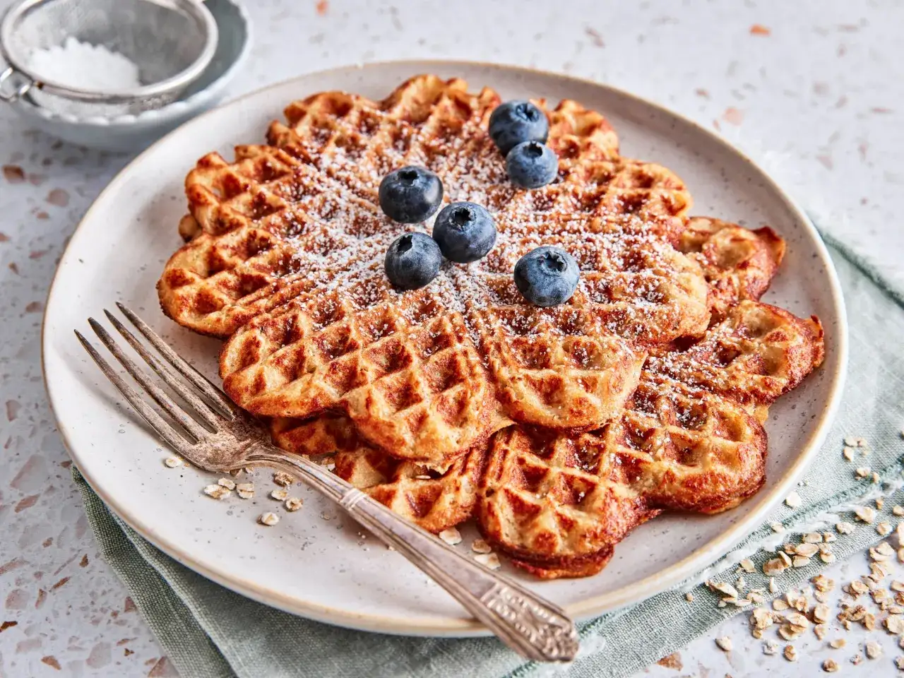 Goldbraune Haferflocken Waffeln, bestäubt mit Puderzucker und garniert mit frischen Blaubeeren, auf einem Teller mit einer Gabel.