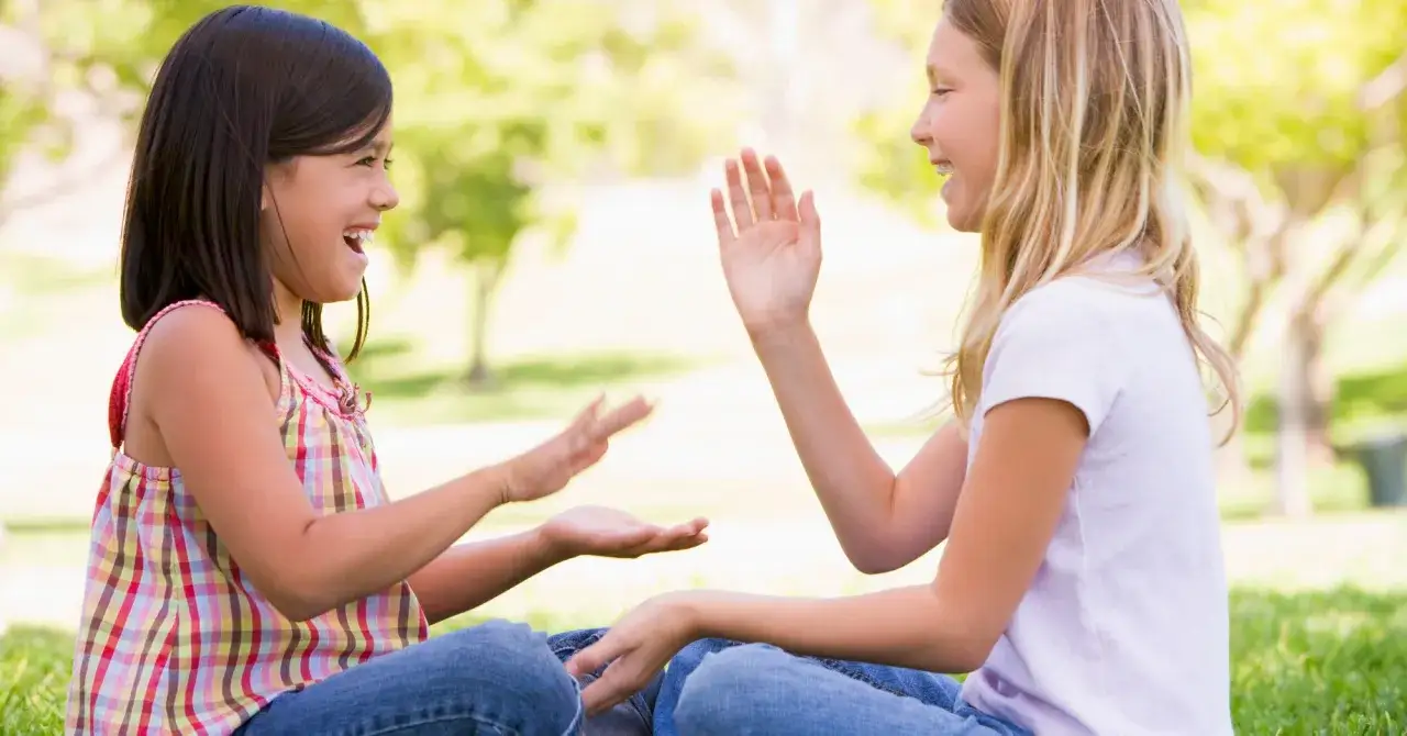 Dos ni&ntilde;as r&iacute;en mientras juegan a los juegos de palmas sentadas en el c&eacute;sped bajo los &aacute;rboles.