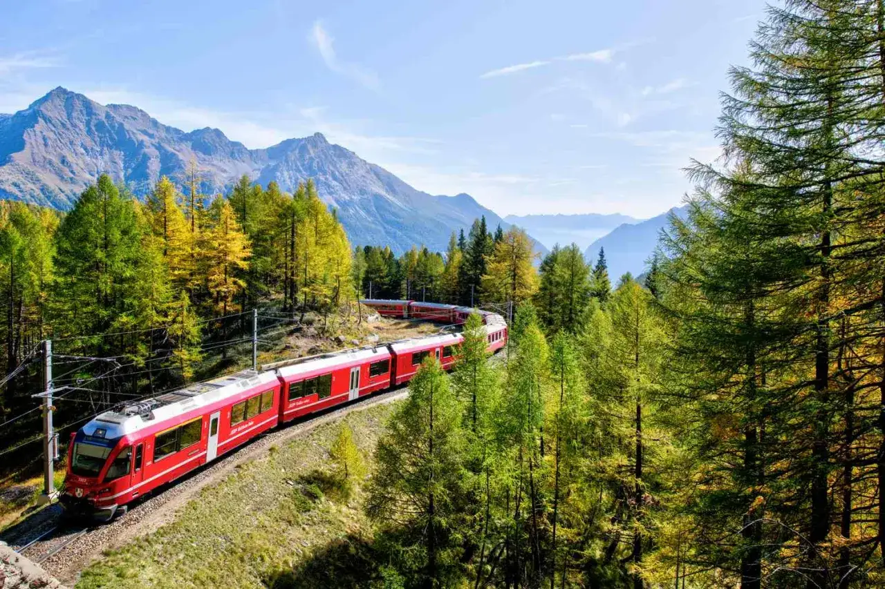 Roter Zug schl&auml;ngelt sich durch eine Berglandschaft mit bunten B&auml;umen. So funktioniert Interrail: Freiheit auf Schienen durch atemberaubende Panoramen.