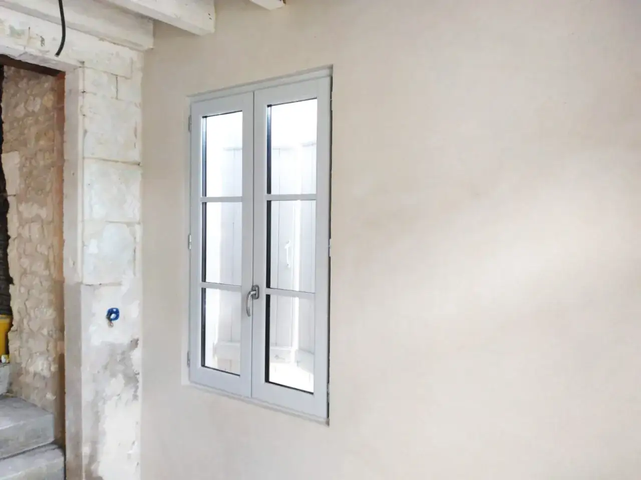 Un mur int&eacute;rieur avec un enduit chaux li&egrave;ge, une fen&ecirc;tre blanche &agrave; double battant et une partie d'un mur en pierre brute.