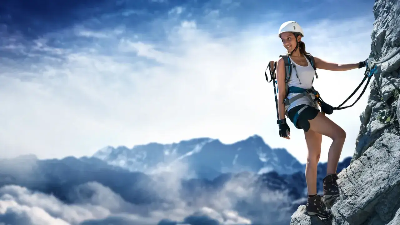 Kobieta na via ferrata polska, w kasku i uprzęży, wspina się po skalnej ścianie. W tle góry i chmury.