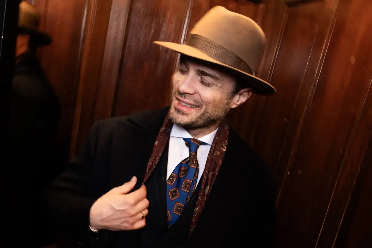 Mężczyzna w brązowym kapeluszu gangstera, czarnym płaszczu i eleganckim krawacie.