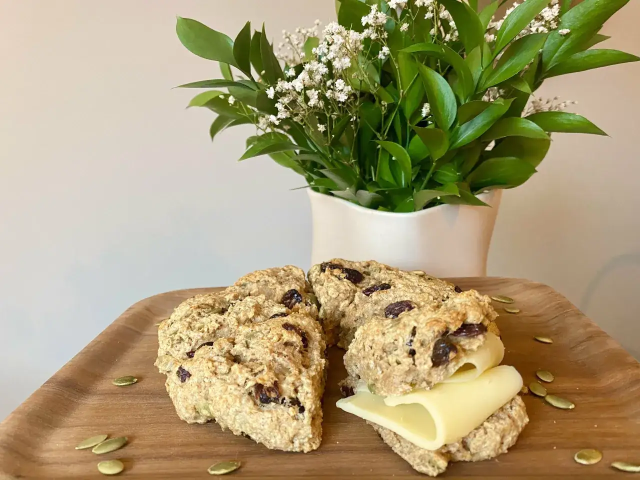 Grova scones med russin och pumpafrön, en med ost, på en träbricka. En bukett med gröna blad och små vita blommor står bakom.