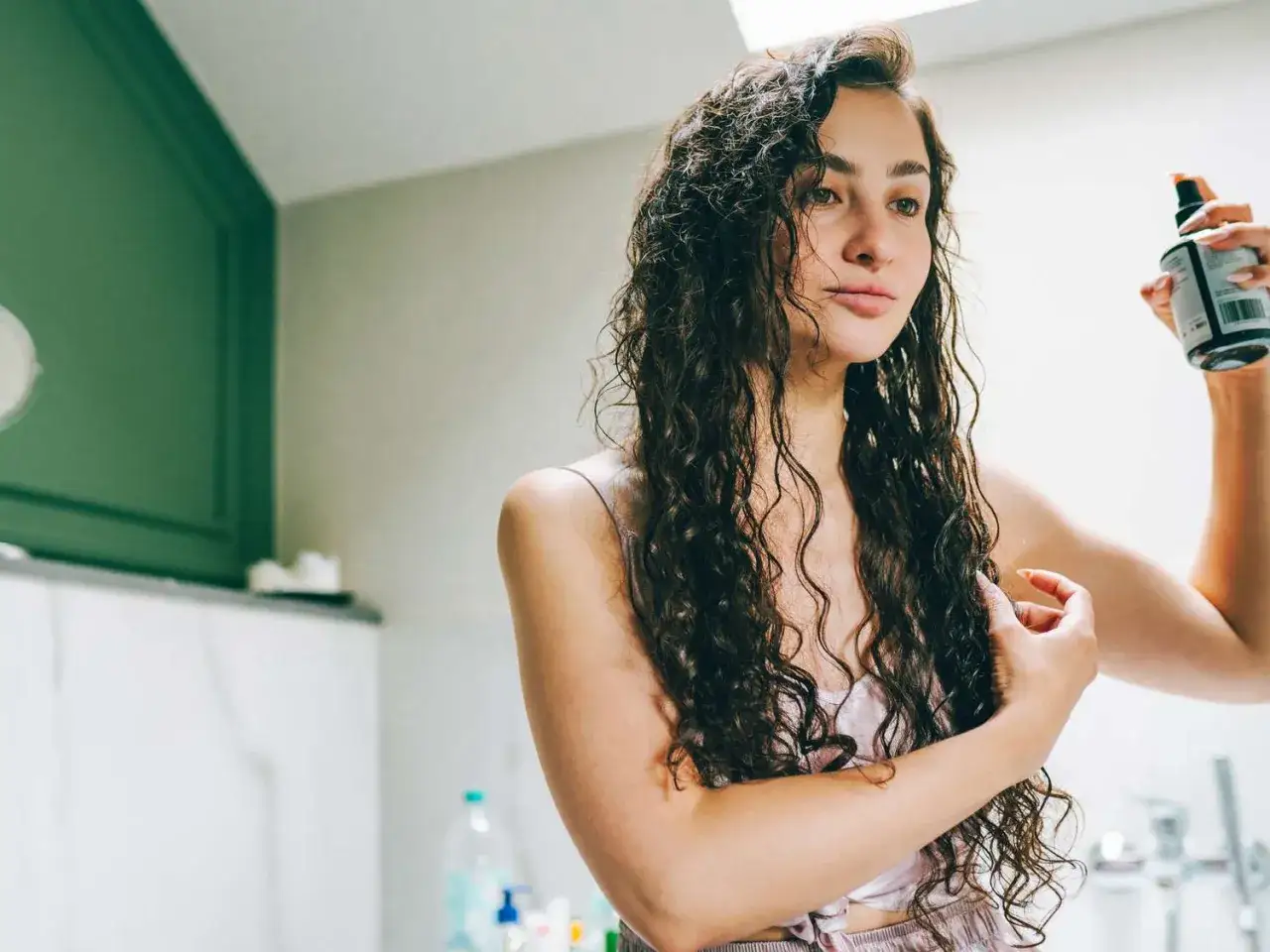 Frau mit langen, lockigen Haaren sprüht Olaplex Nr. 6 auf ihr feuchtes Haar, um es zu pflegen und zu stylen.