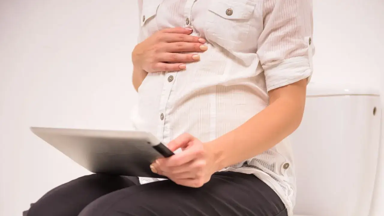 En gravid kvinna sitter på en toalett och håller en surfplatta. Hon har handen på magen, orolig för brunt blod.
