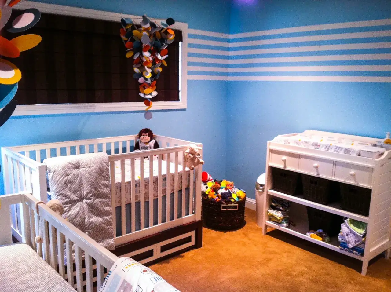 A cozy twin m nursery with two cribs, a changing table, and a basket overflowing with colorful toys.