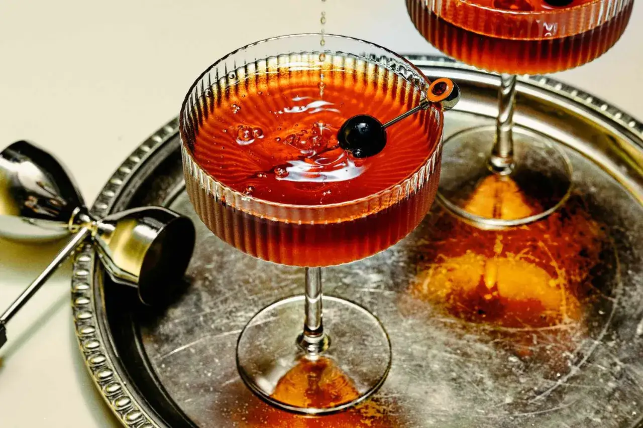 Ein Cocktail mit trockenem Wermut, garniert mit einer Kirsche, auf einem Silbertablett. Daneben ein Jigger.
