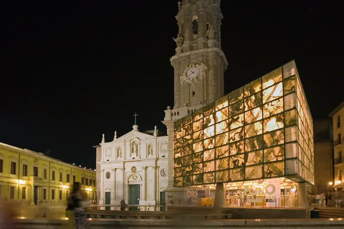 El moderno museo del foro de Caesaraugusta contrasta con la iglesia y su torre medieval iluminados por la noche.