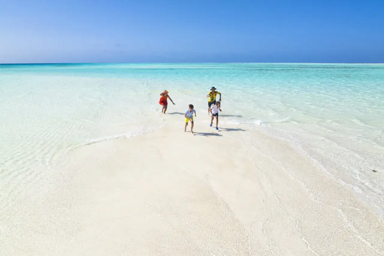 Une famille profite de ses vacances à l'étranger, courant joyeusement sur une plage de sable blanc bordée par une eau turquoise cristalline.