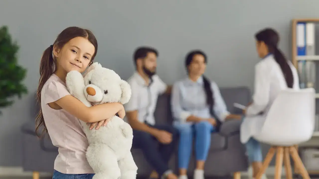 Uśmiechnięte dziecko neurotypowe przytula misia, podczas gdy rodzice rozmawiają z lekarzem w tle.