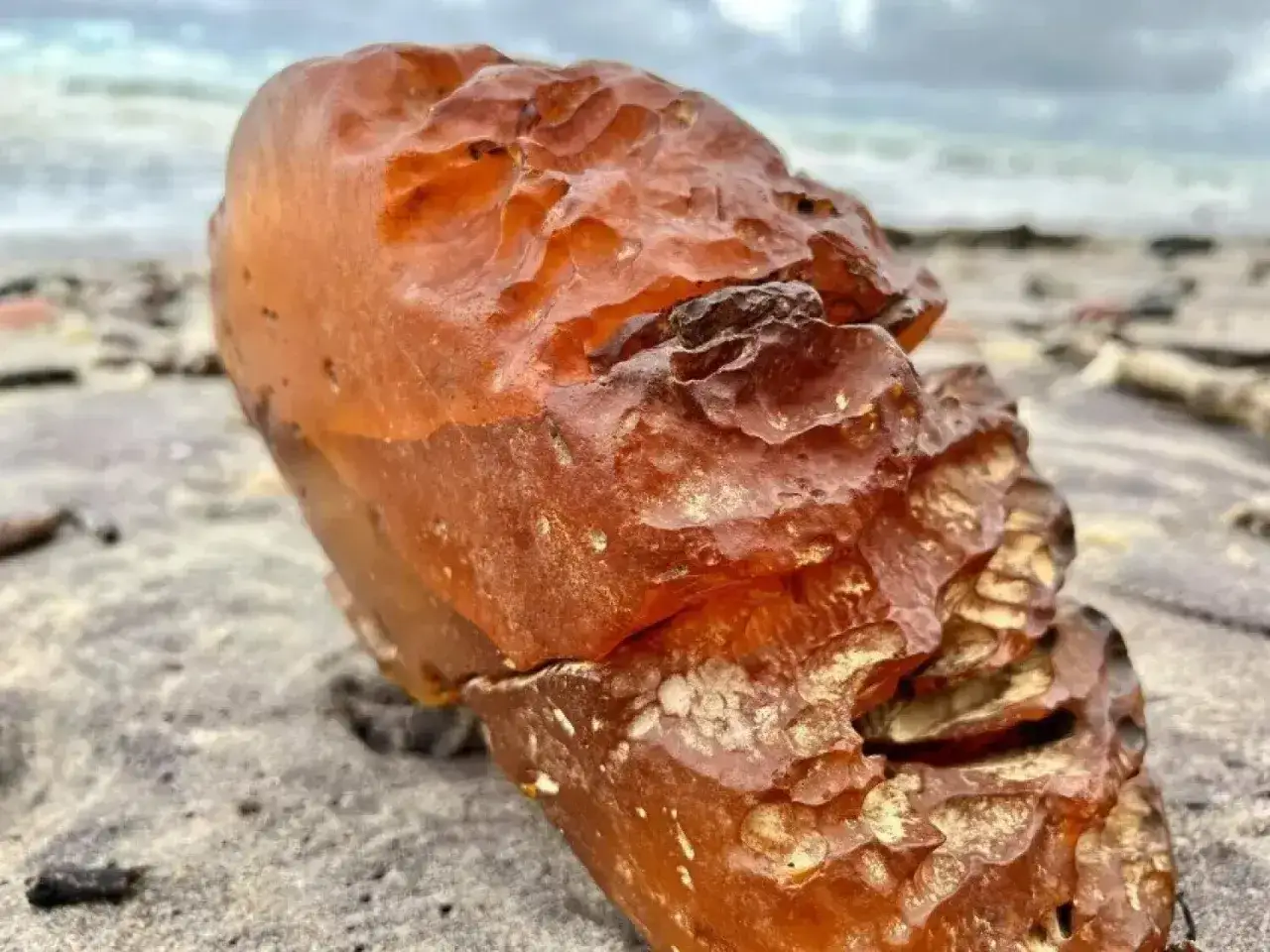 Duży, pomarańczowy bursztyn z widocznymi nierównościami leży na piaszczystej plaży. W tle widać morze i zachmurzone niebo.